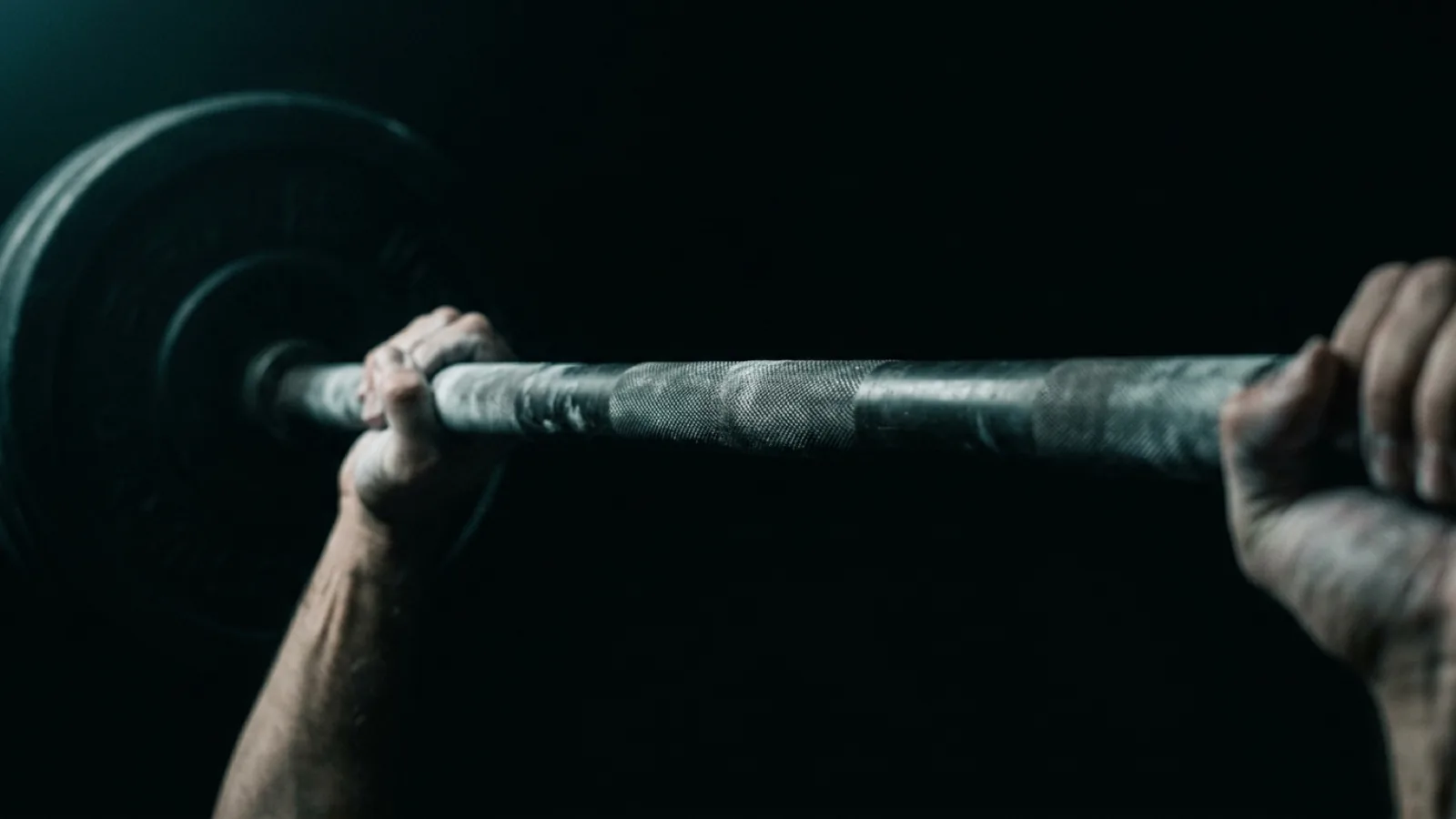 Close-up of a knurled barbell held mid-lift by chalked hands out of frame, the far end of the bar dissolving into darkness.